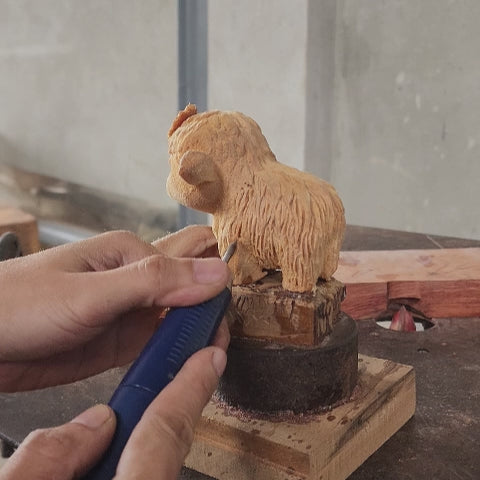 Close-up video showing the artisan hand-carving process of a BeeRusty wooden Highland Cow figurine. The artist uses tools to create detailed fur texture on the solid wood sculpture, proving authentic craftsmanship